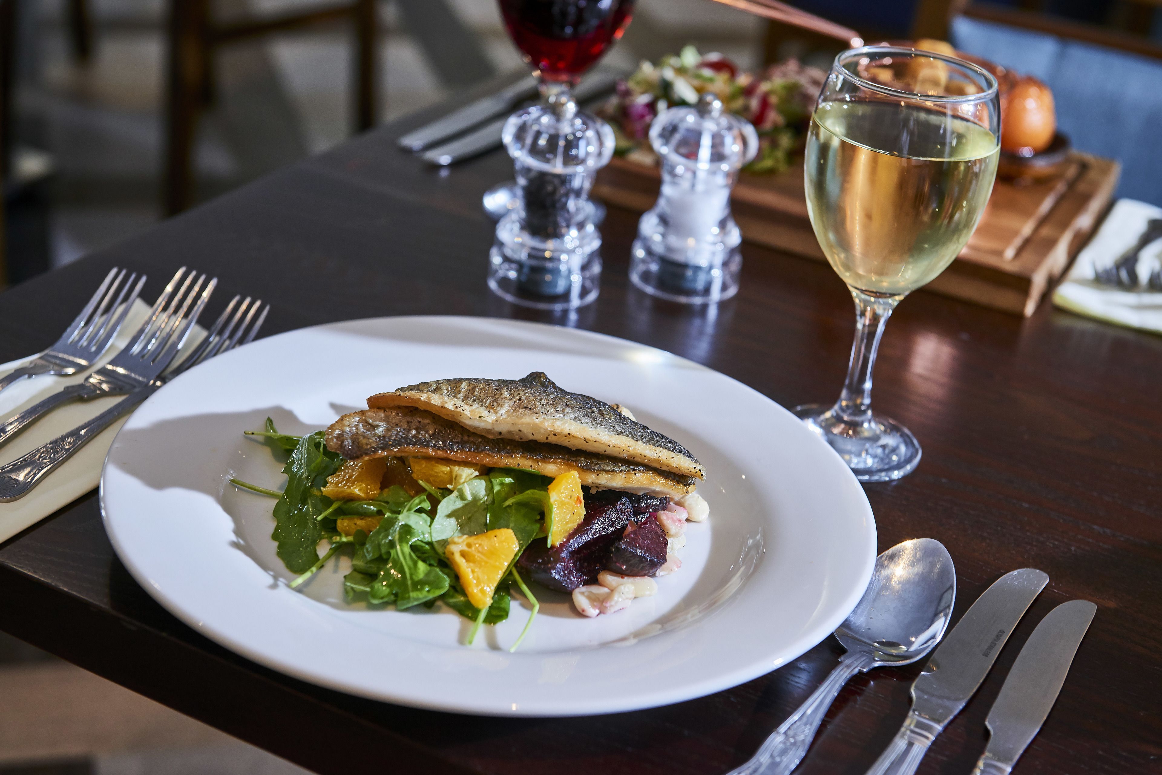 Un plato de comida con pescado, ensalada y una copa de vino blanco sobre una mesa de madera.