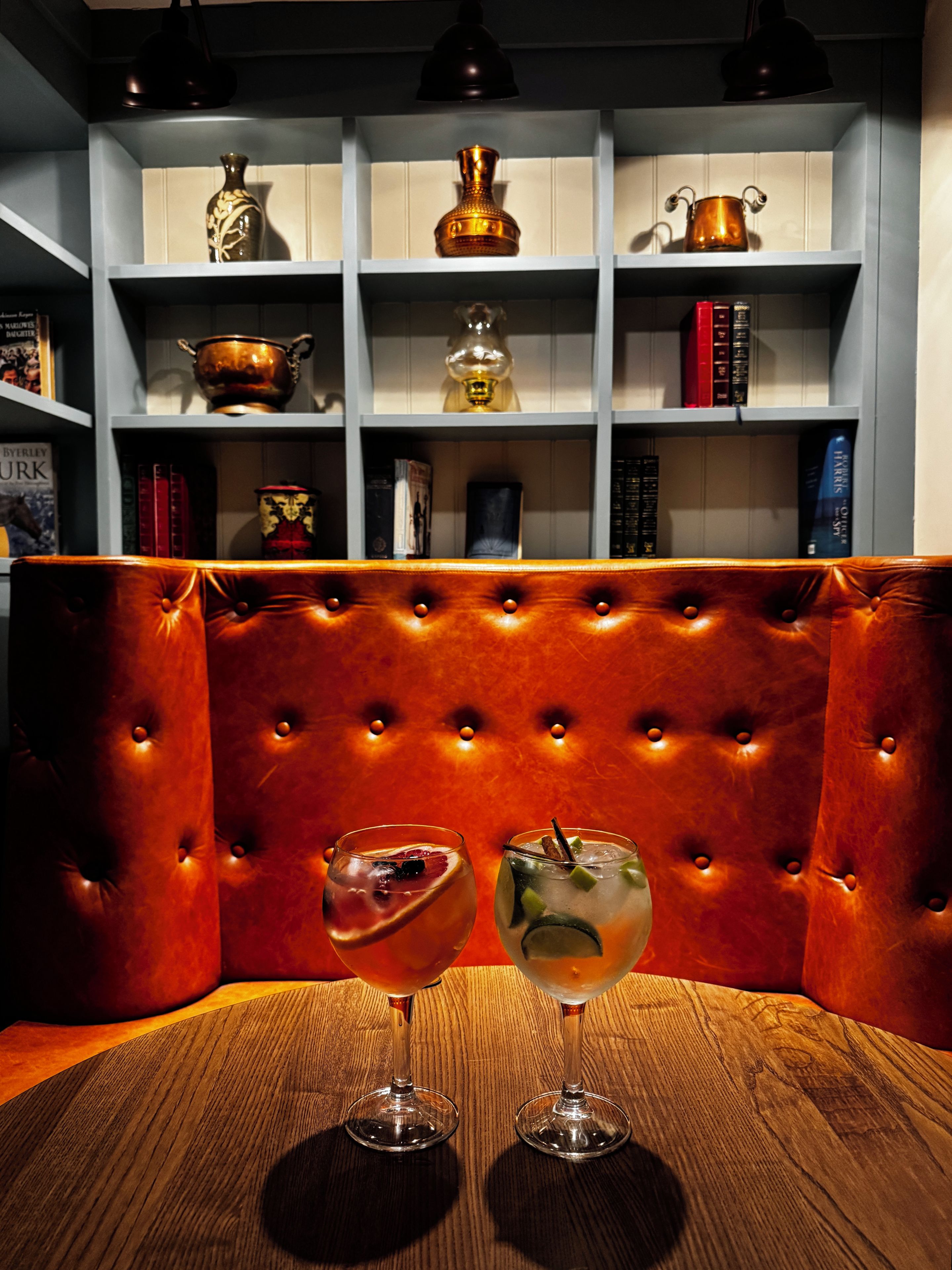 A room with a wooden table, two glasses of drinks, a bookshelf with various items, and a tufted leather bench.