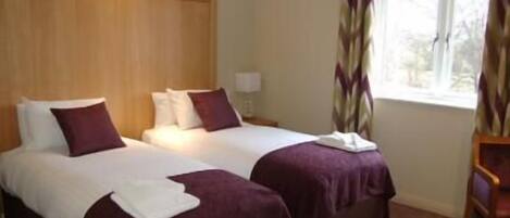 Habitación de hotel con dos camas, cada una con colcha blanca y morada, cabecero de madera y ventana con cortinas.
