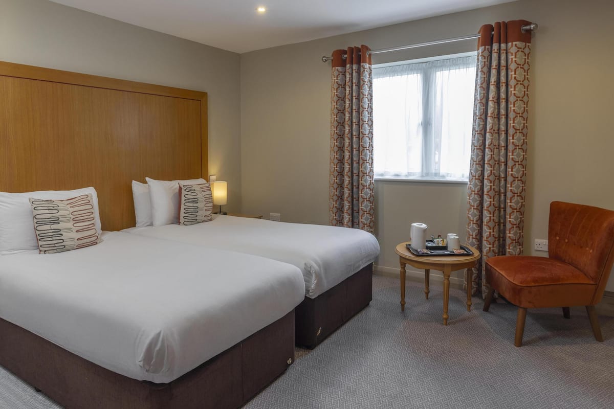 A hotel room with two beds, a wooden headboard, a small round table with toiletries, and a single chair.