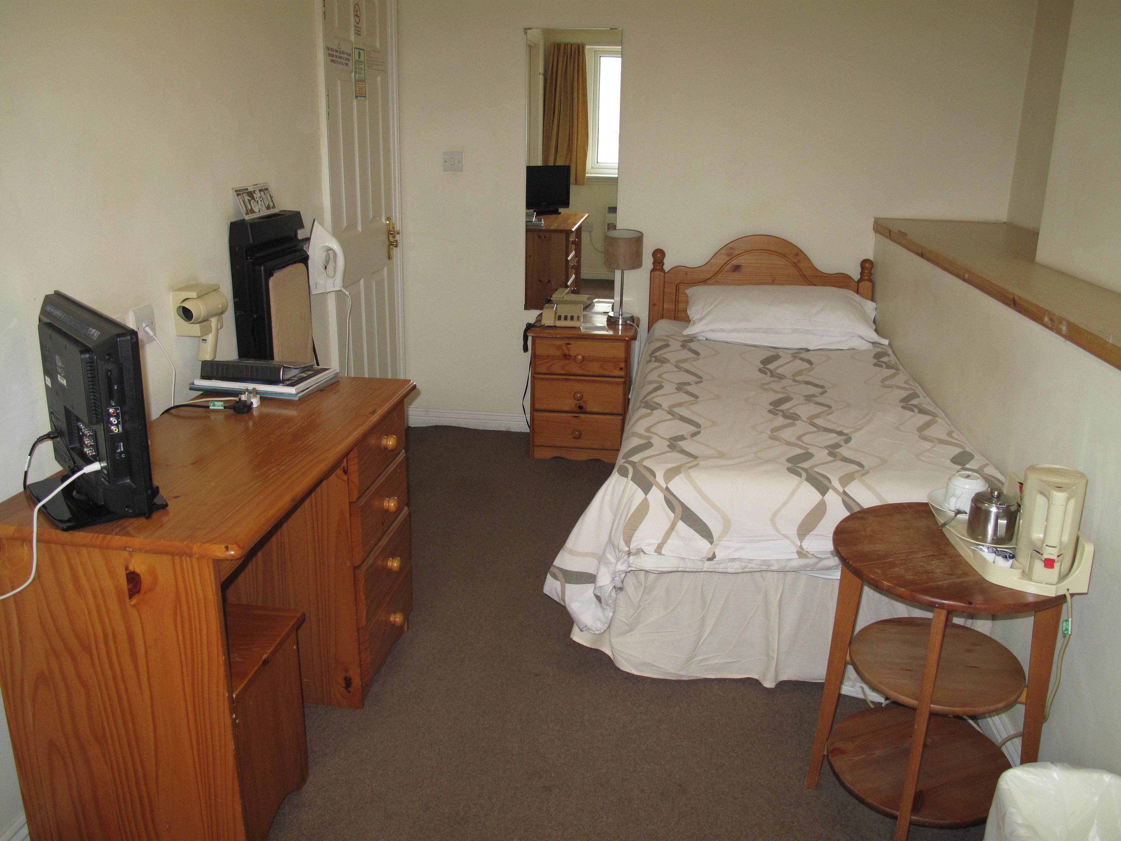A bedroom with a bed, a desk with a computer, a dresser, and a bedside table with a lamp and a cup.