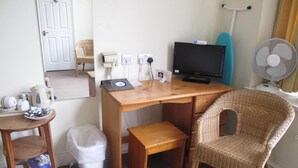 Une pièce de taille modeste, comprenant un bureau en bois, une chaise en osier, un téléviseur, une petite table avec une bouilloire et des tasses, ainsi qu’une poubelle blanche.