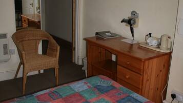 Un dormitorio con una cama, un aparador de madera, una silla de mimbre y un escritorio con una computadora y una cafetera.