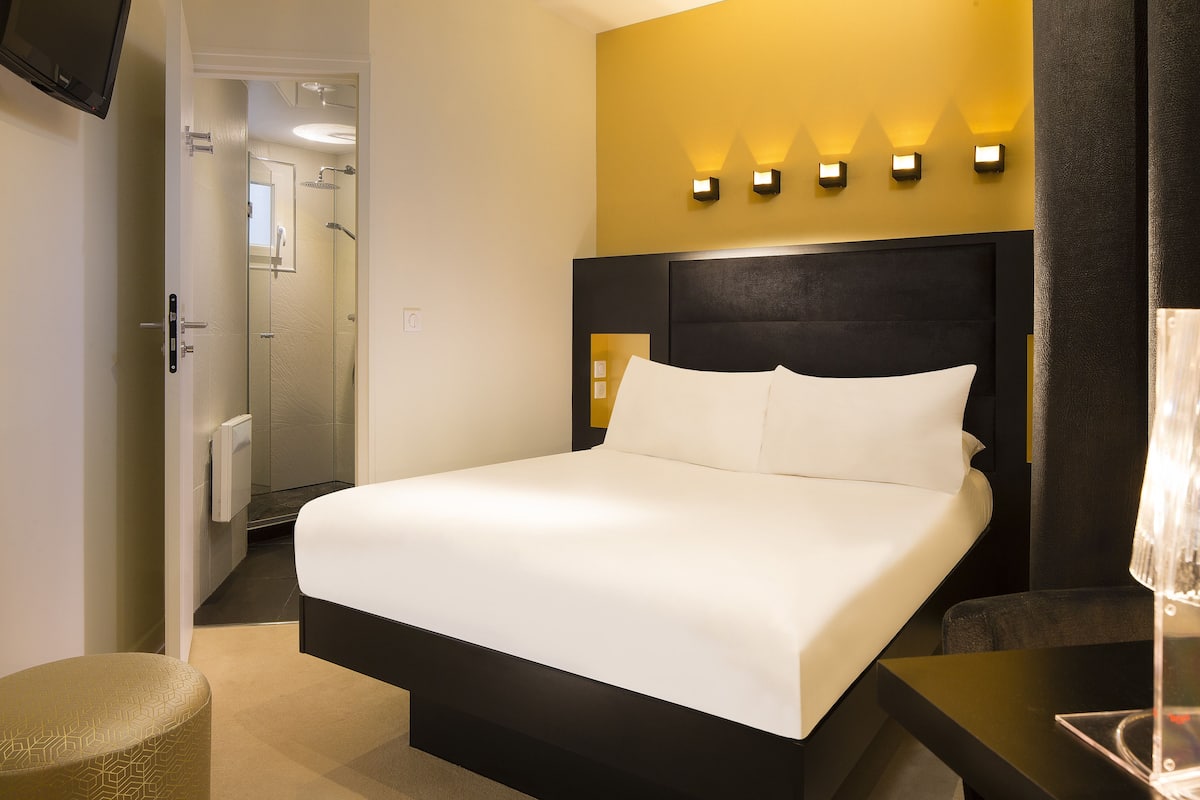A hotel room with a large bed, a bedside table, a nightstand, a wall-mounted TV, and a bathroom visible through an open door.