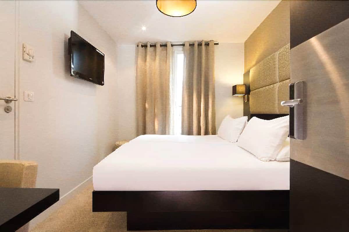 A hotel room with a bed, a TV mounted on the wall, a bedside table, and a window with curtains.