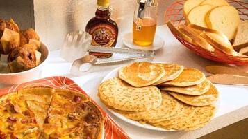 Une table de petit-déjeuner avec divers aliments, notamment des crêpes, une bouteille de sirop et un panier de pain.