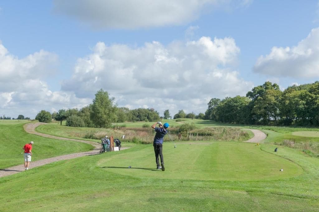 Campo de golf con cielo despejado, un golfista en pleno balanceo y un camino que lleva a un tee.