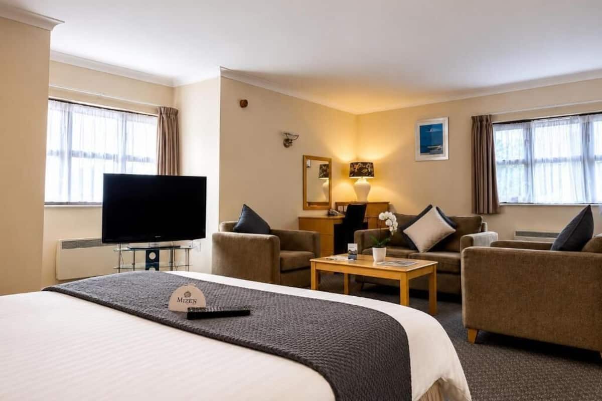 A hotel room with a bed, a television, a desk with a lamp, two armchairs, and a coffee table.