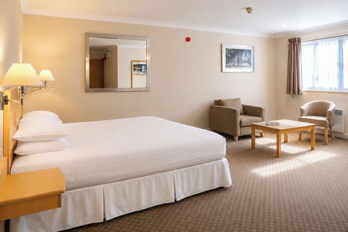 A hotel room with a large bed, a desk, a chair, a small table, a mirror, and a window with curtains.
