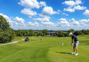Um campo de golfe sob um céu claro, um jogador se preparando para dar a tacada final e um carrinho de golfe ao fundo.