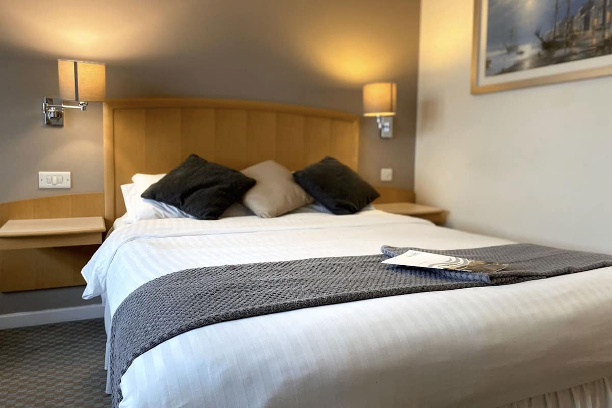 A hotel room with a bed, two bedside lamps, a nightstand, a painting on the wall, and a power outlet.