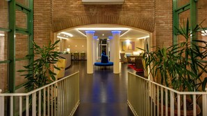 A hotel lobby with a brick wall, a reception desk, a blue seating area, and potted plants. - Hotel San Marco (Lucca)