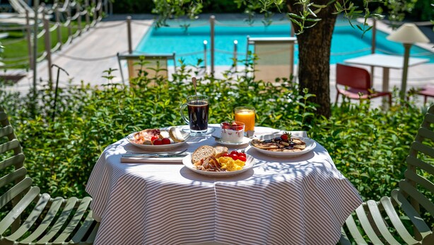 A dining table with breakfast items, a pool in the background, and greenery around. - Hotel San Marco (Lucca)