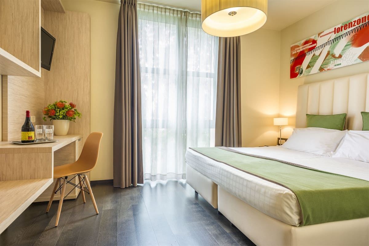A hotel room with a bed, a desk with a chair, a TV mounted on the wall, and a window with curtains.