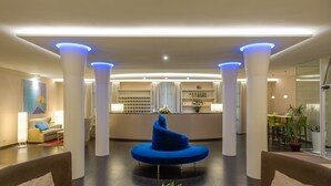 A modern lobby with a central blue seating area, white pillars, and a reception desk in the background. - Hotel San Marco (Lucca)