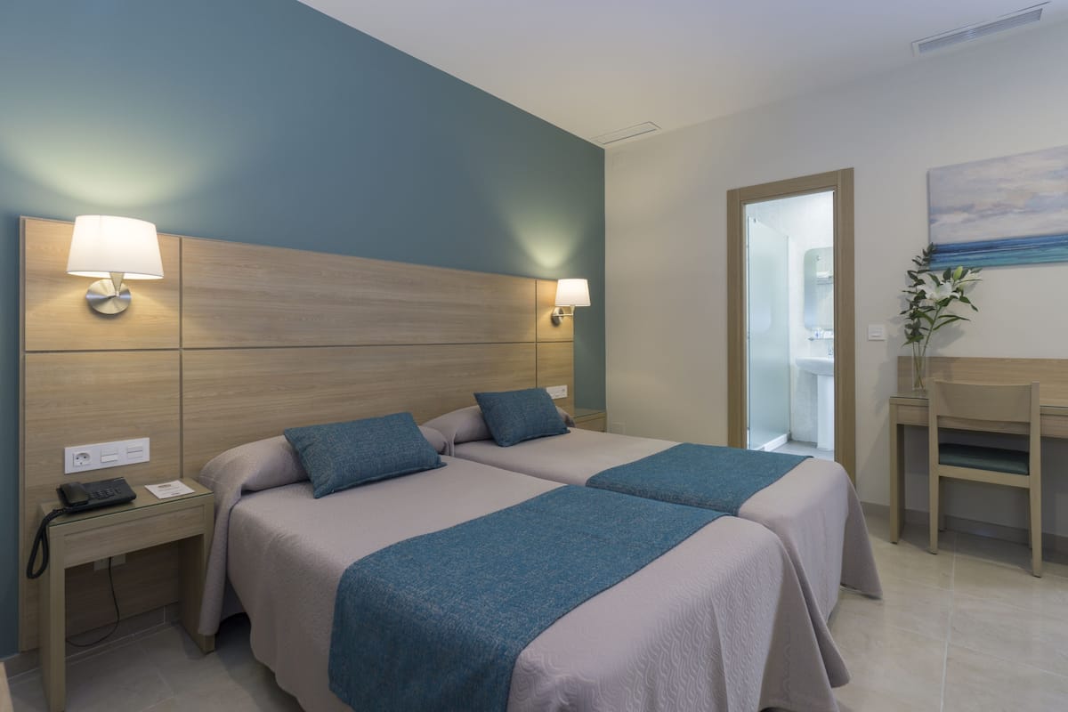 Habitación de hotel con dos camas, un escritorio y una silla. Hay lámparas de noche, un teléfono y una mesita con una lámpara.