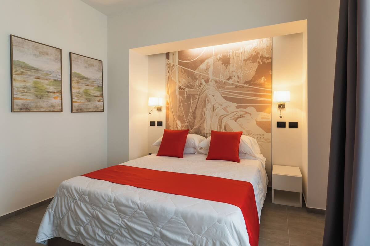 A hotel room with a large bed, two red pillows, a white bedspread, two wall-mounted lamps, and two framed artworks on the wall.