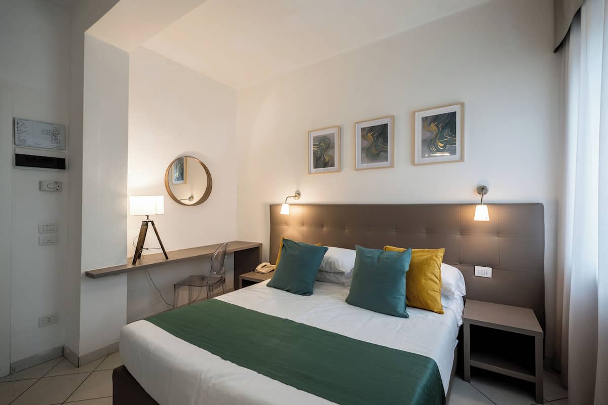 A hotel room with a neatly made bed, a bedside table, a desk with a lamp, a chair, and artwork on the wall.