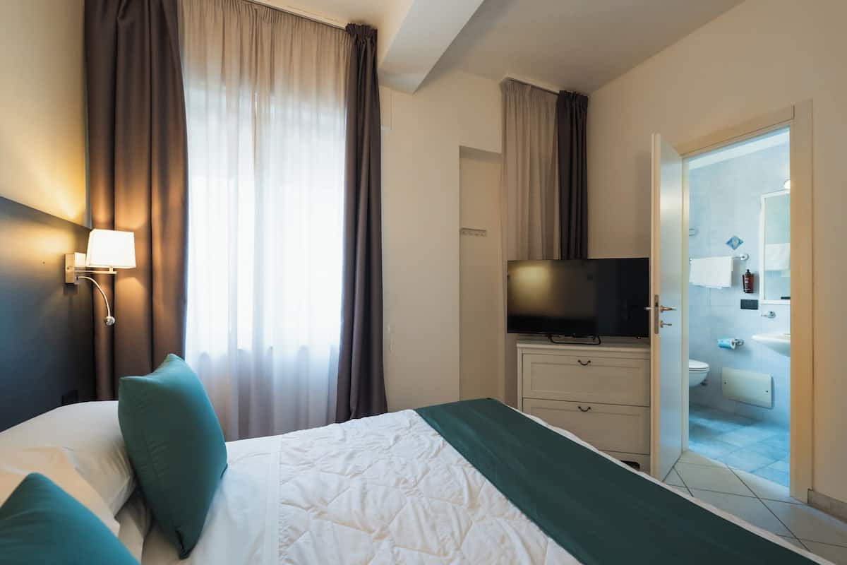 A hotel room with a bed, a TV, a bathroom visible through an open door, and a window with curtains.