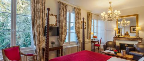 A hotel room with a large bed, a red chair, a fireplace, and a view of trees through the window.