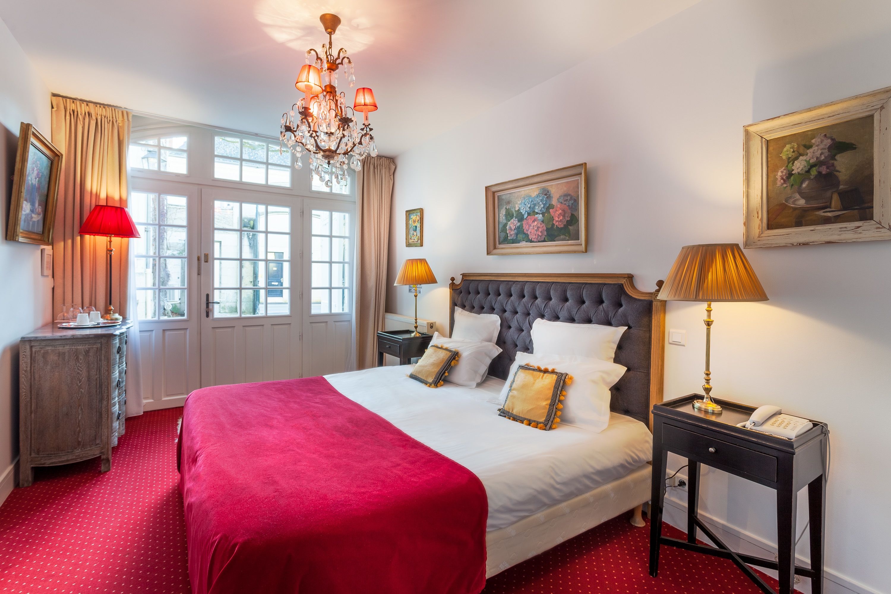 A hotel room with a large bed, a chandelier, a nightstand with a lamp, and a painting on the wall.
