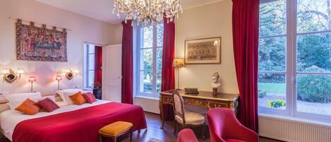 Una habitación de hotel con una cama grande, un escritorio, una silla, un sillón rojo, una araña y ventanas amplias con cortinas.