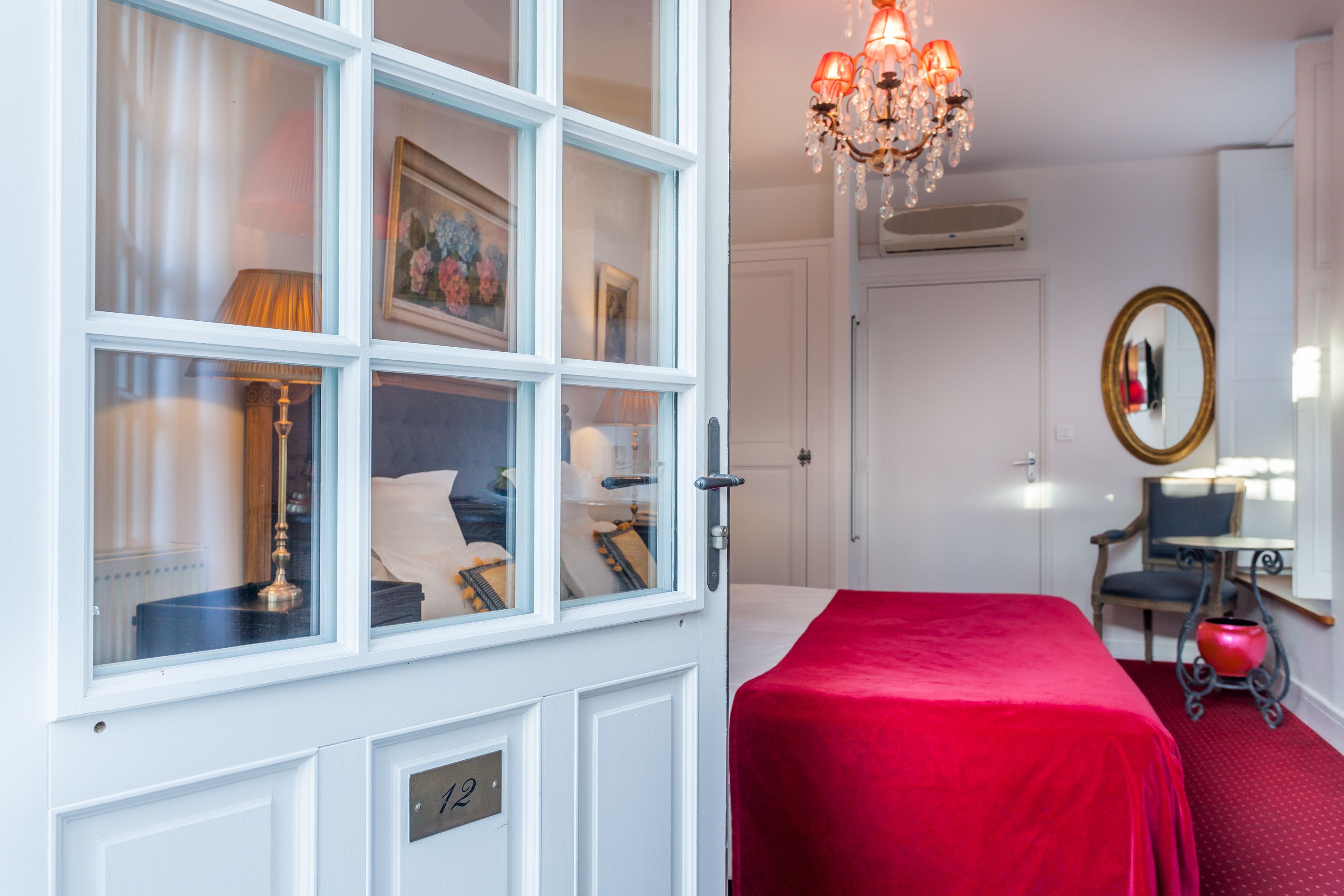Een hotelkamer met een groot raam, een kroonluchter, een bed met een rode sprei, een bureau met een lamp en een spiegel aan de muur.