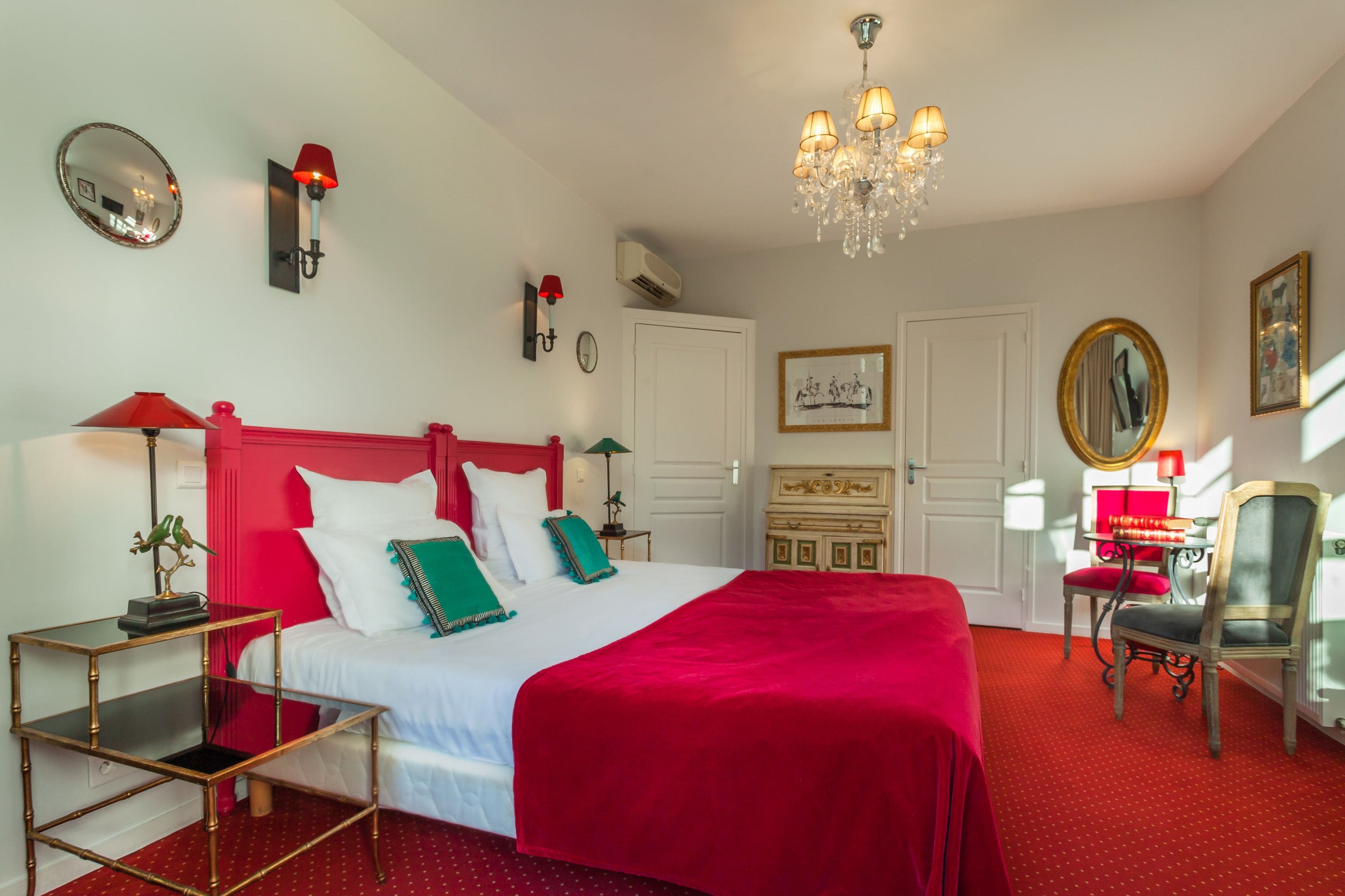 A hotel room with a large bed, red bedding, a chandelier, a red-framed mirror, and a red armchair.