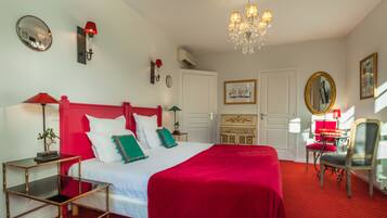 A hotel room with a large bed, red bedding, a chandelier, a red-framed mirror, and a red armchair.