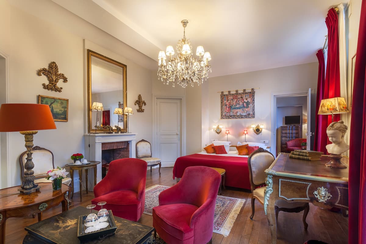 A room with a bed, two red armchairs, a wooden desk, a chandelier, and a fireplace.