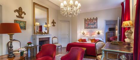A room with a bed, two red armchairs, a wooden desk, a chandelier, and a fireplace.