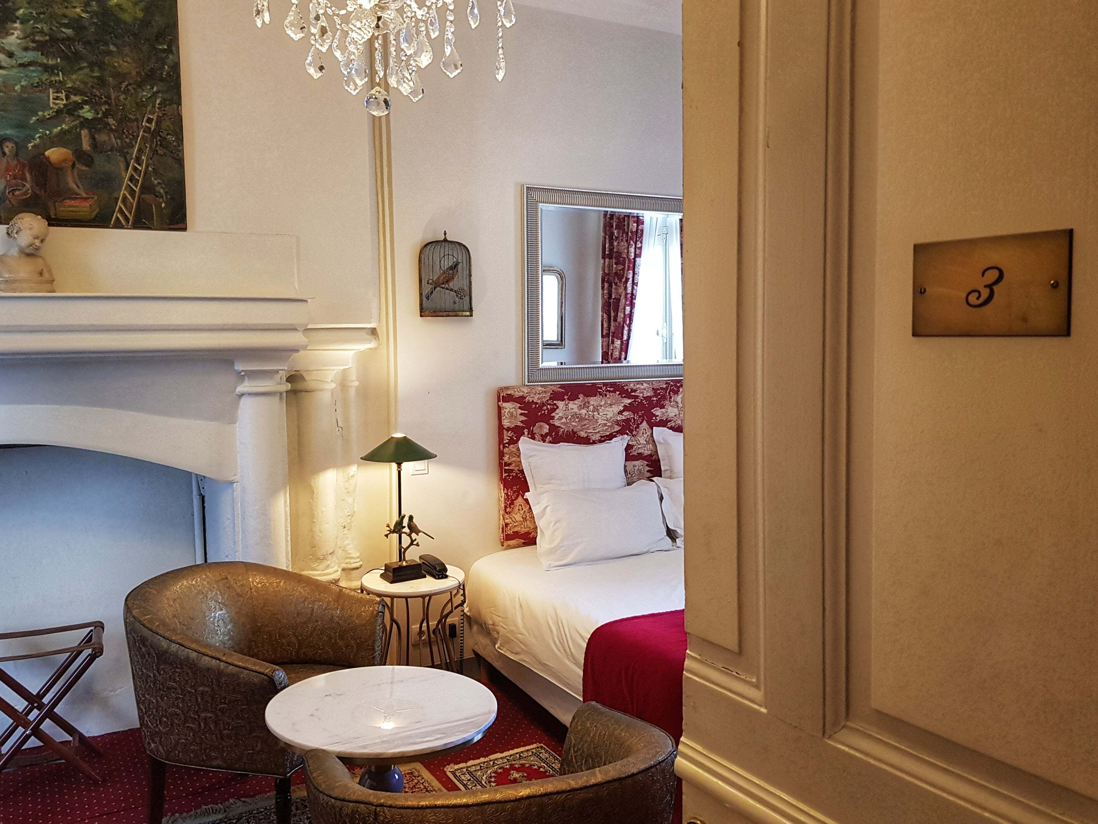 A hotel room with a fireplace, a bed with a red headboard, a chandelier, and a small table with a lamp.