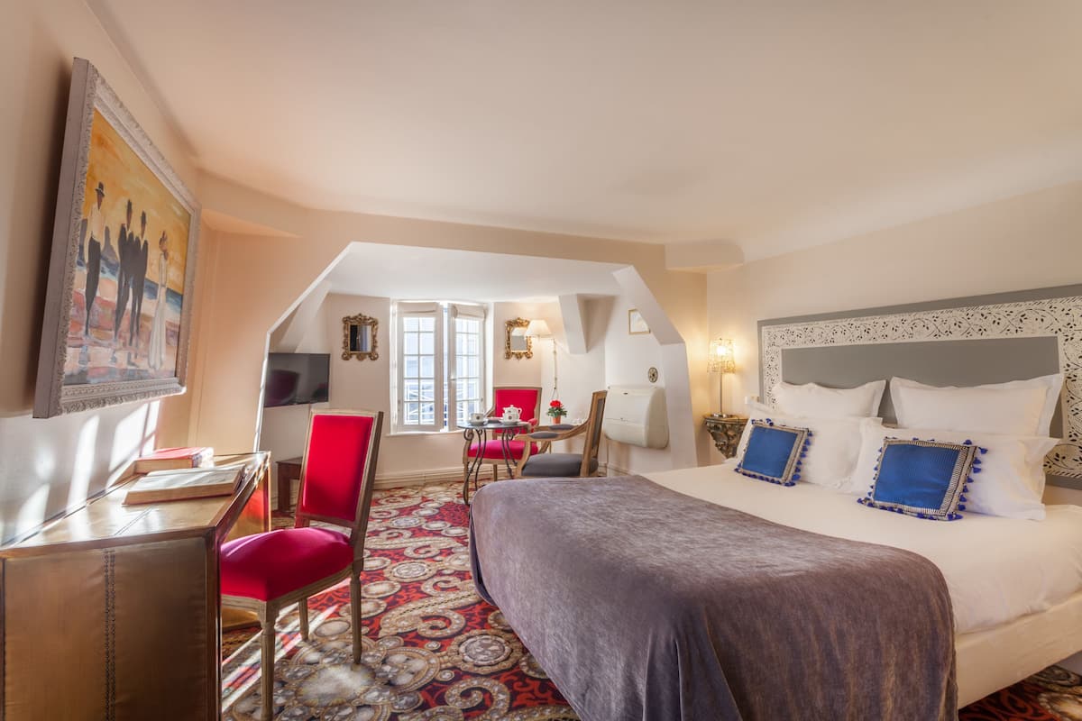 A hotel room with a large bed, a desk, a chair, and a red chair. There is a painting on the wall and a window with curtains.