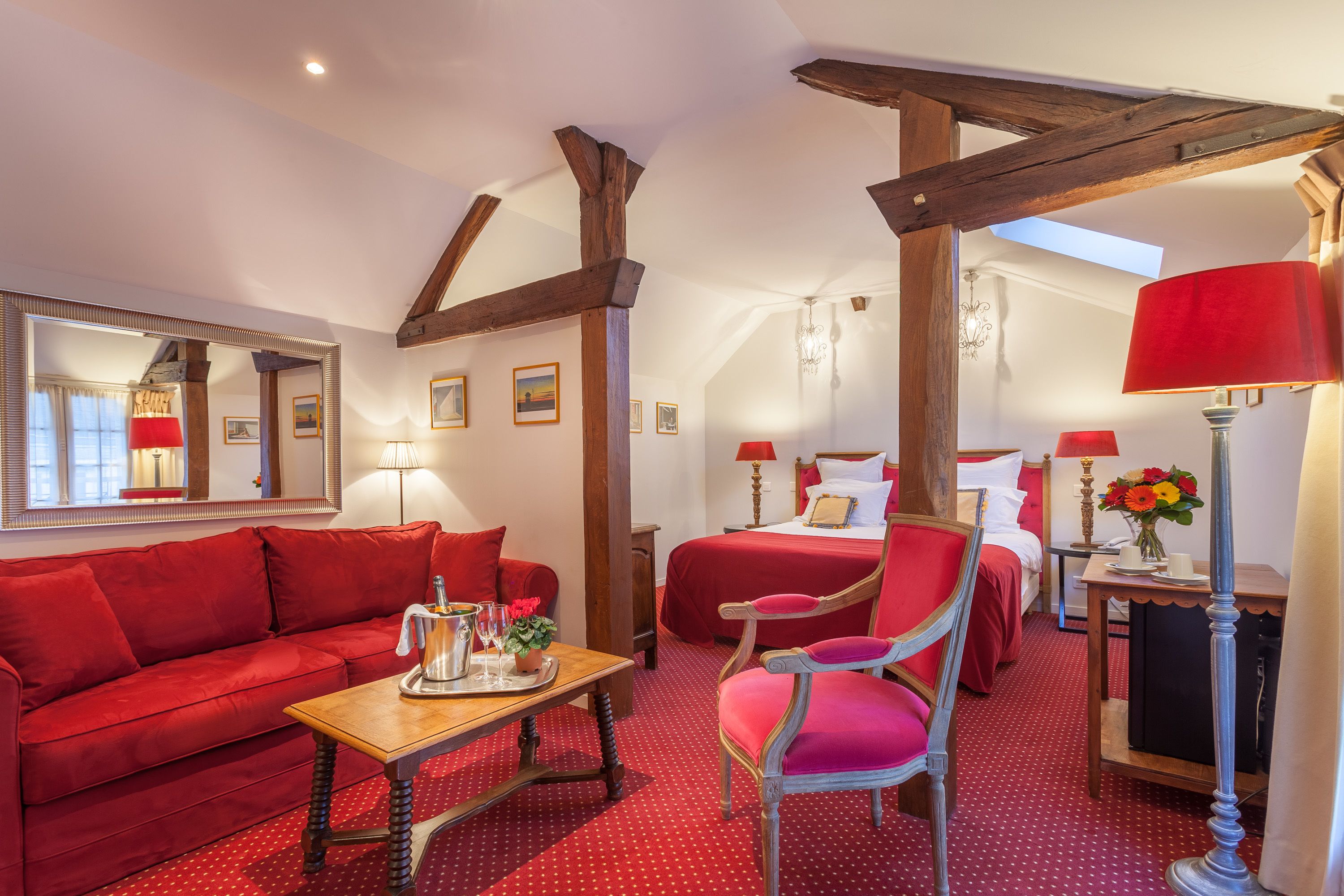 Une chambre d’hôtel avec un canapé rouge, une table en bois, un fauteuil rouge, un lit avec une literie rouge et un miroir au mur.