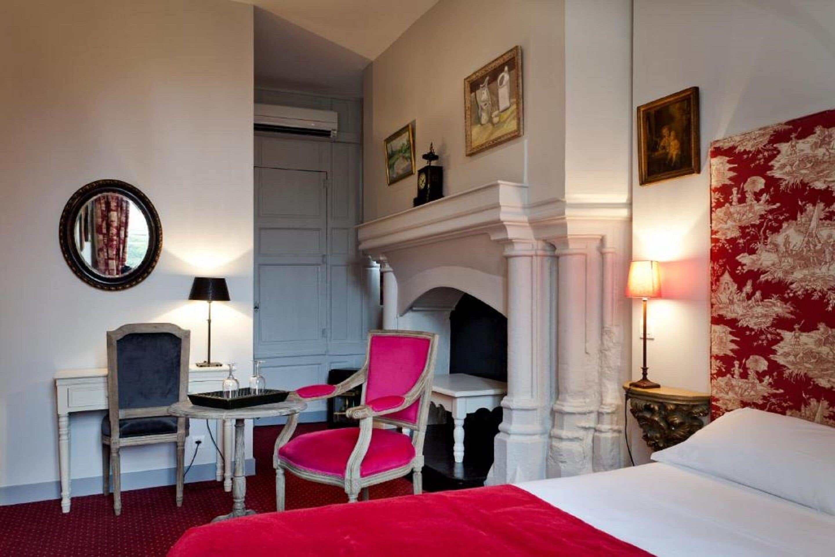 A hotel room with a bed, two chairs, a small table, a fireplace, and a mirror on the wall.