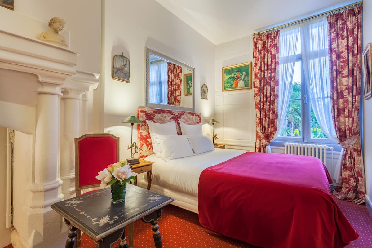 A bedroom with a large bed, a red chair, a small table with a vase, and a window with curtains.