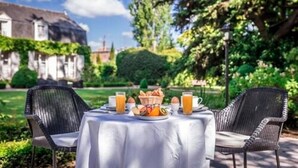 Una mesa de comedor al aire libre con alimentos para el desayuno, rodeada de sillas de mimbre, con un jardín y un edificio al fondo.
