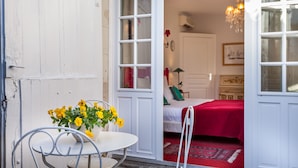 Una habitación con una cama, una colcha roja, una puerta blanca y un ventanal con candelabro.