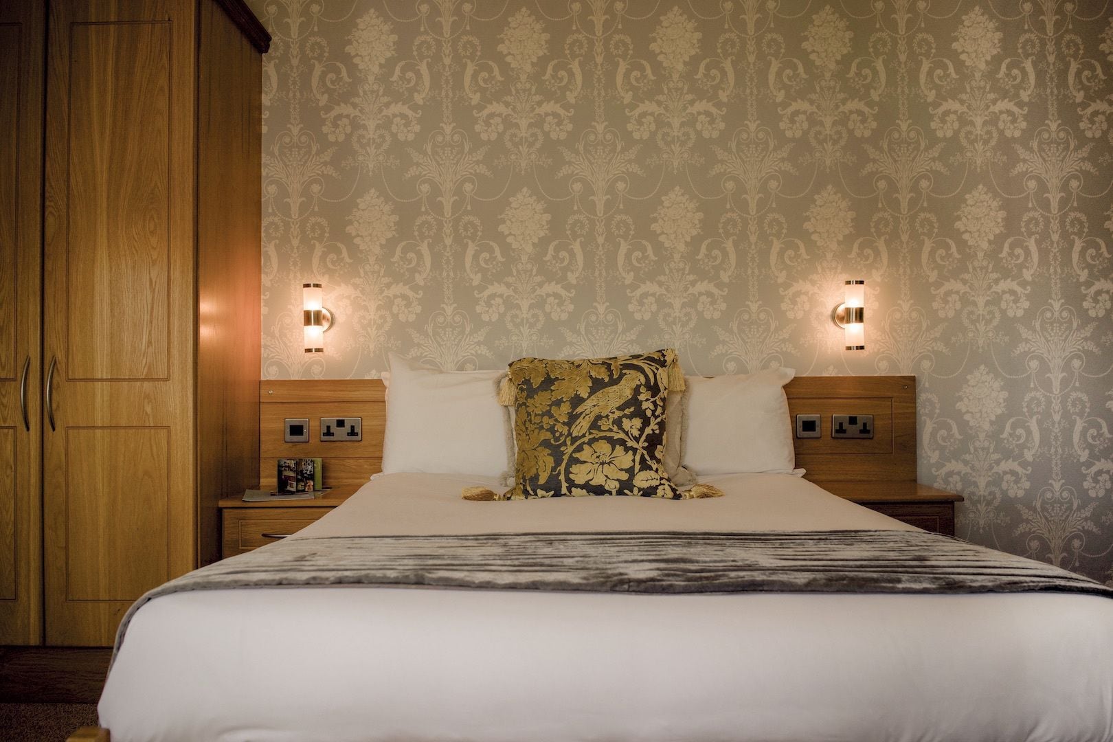 Una habitación de hotel con una cama grande, dos mesitas de noche, un armario de madera y papel tapiz estampado.