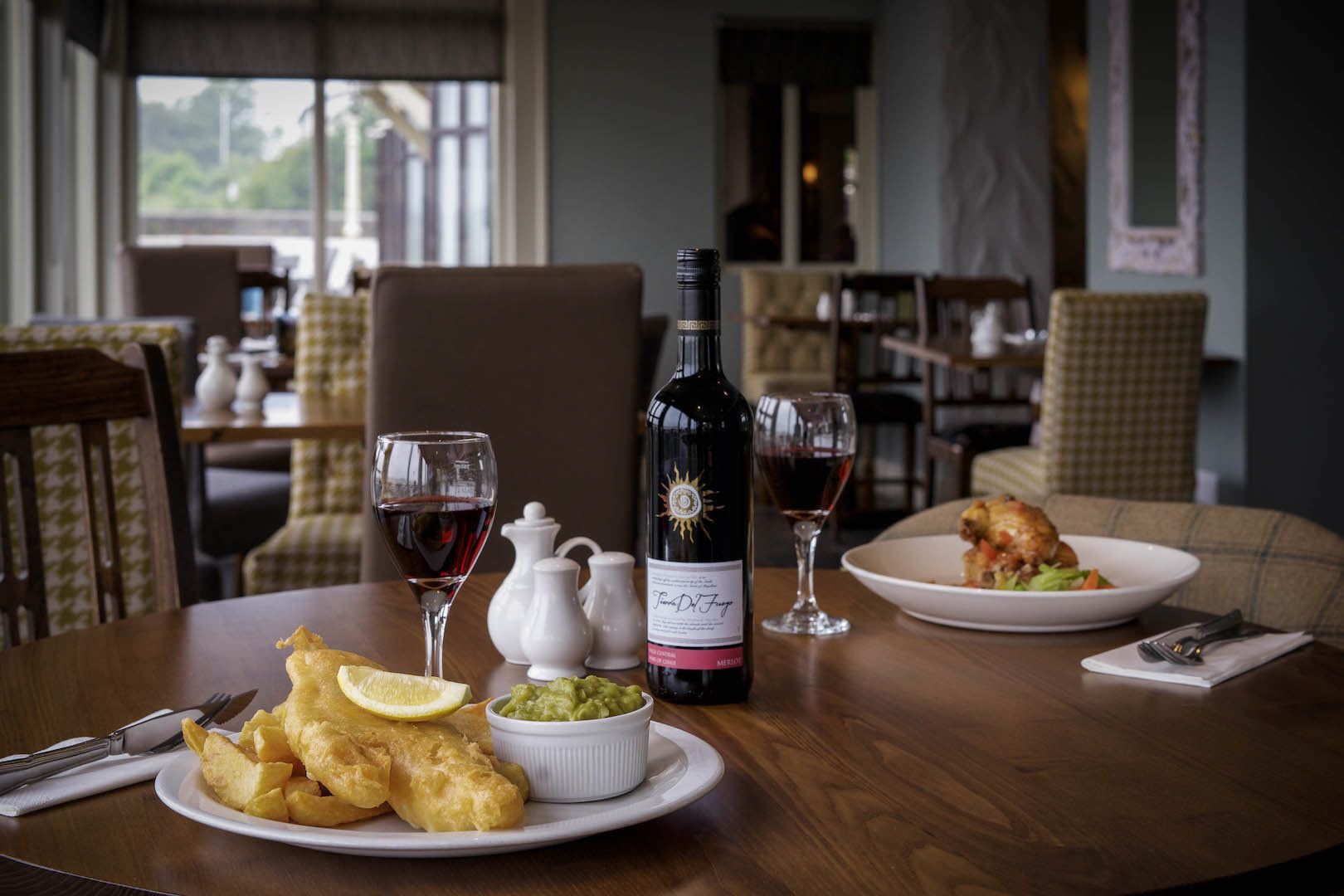 A dining table with a plate of fish and chips, a bottle of wine, and a glass of red wine.