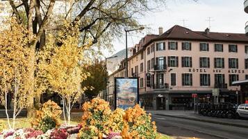 Un hôtel à la façade de style européen classique, avec une enseigne bien visible, situé dans une rue bordée de voitures garées et agrémentée d’un parterre de fleurs.