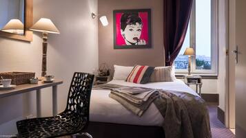 A hotel room with a bed, a chair, a desk with a lamp, a picture on the wall, and a window with curtains.