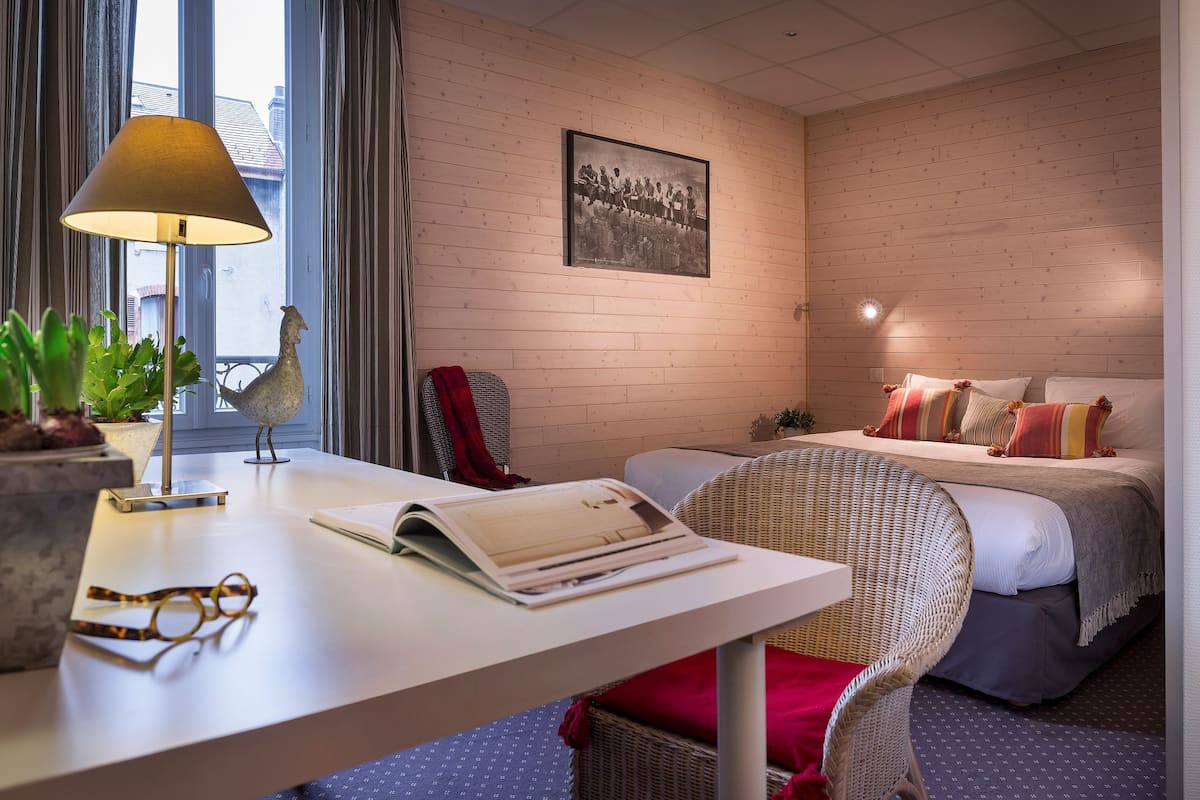 Ett hotellrum med en väggpanel av trä, en säng med kuddar, en fåtölj av rotting, ett skrivbord med en lampa och en bokhylla.