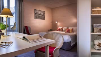 A hotel room with a wooden wall, a bed with pillows, a wicker chair, a desk with a lamp, and a bookshelf.