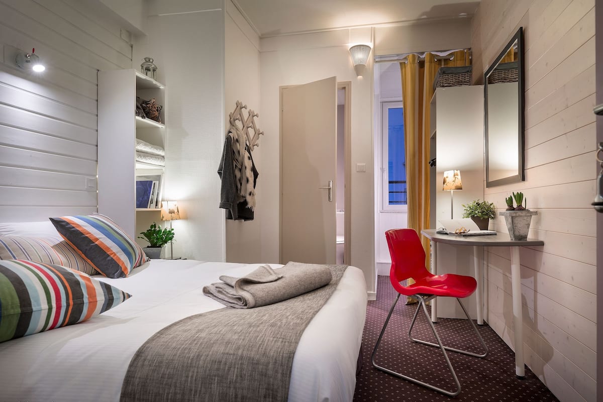 Ett hotellrum med en säng, en röd stol, ett sängbord med en lampa, en spegel och en liten krukväxt.