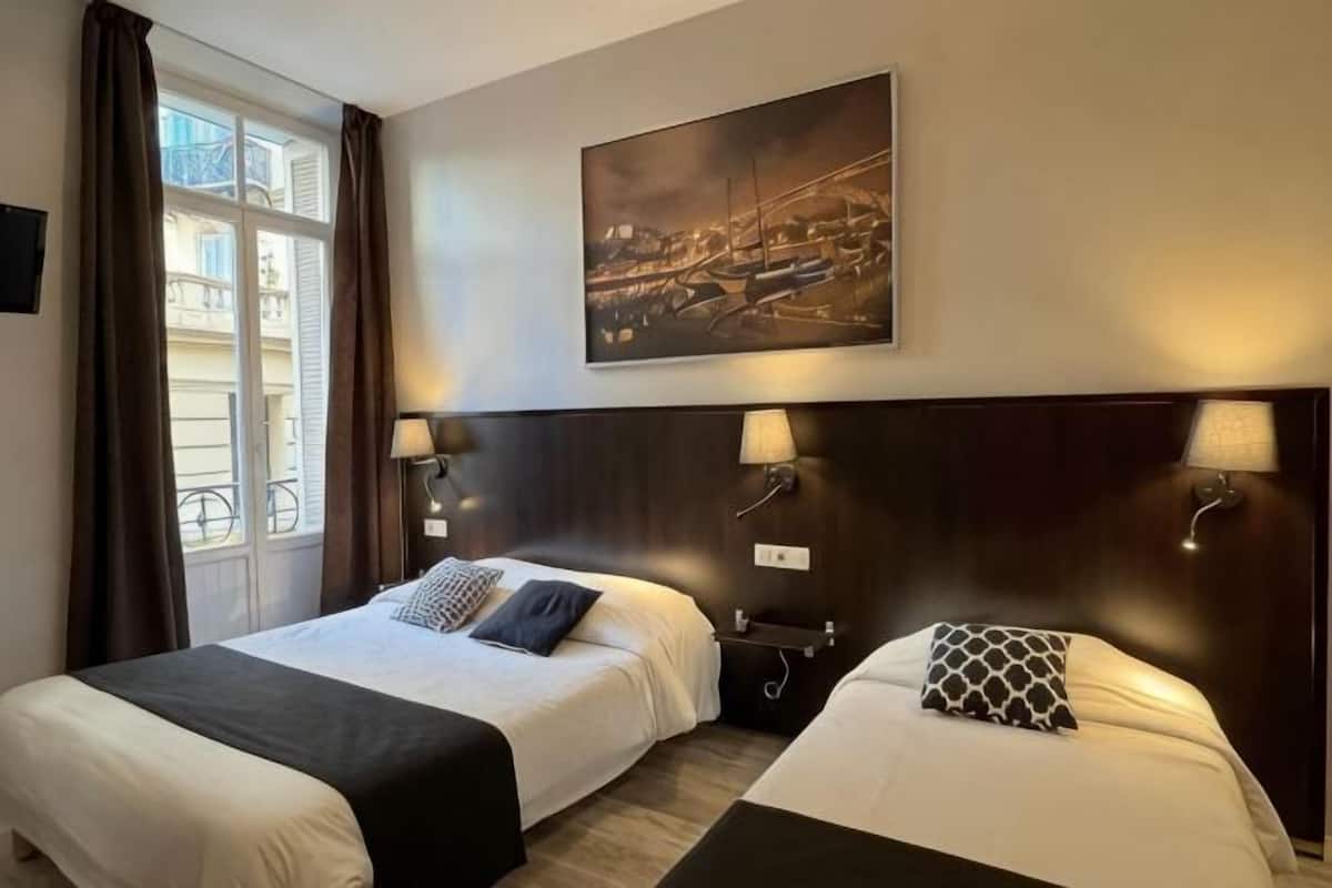 Habitació d'hotel amb dos llits, una finestra gran amb cortines, un quadre de paisatge urbà i làmpades penjades a la paret.