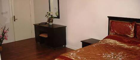 A bedroom with a wooden floor, a bed with a red and gold bedspread, a small desk with a chair, a mirror, and a vase with flowers.