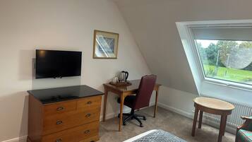 A room with a TV mounted on the wall, a desk with a chair, a dresser, a small round table, and a window with a view of greenery.