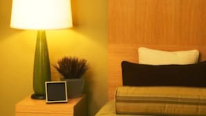Ein Zimmer mit einem Bett, einem Nachttisch mit Lampe, einer kleinen Topfpflanze und einer digitalen Uhr.