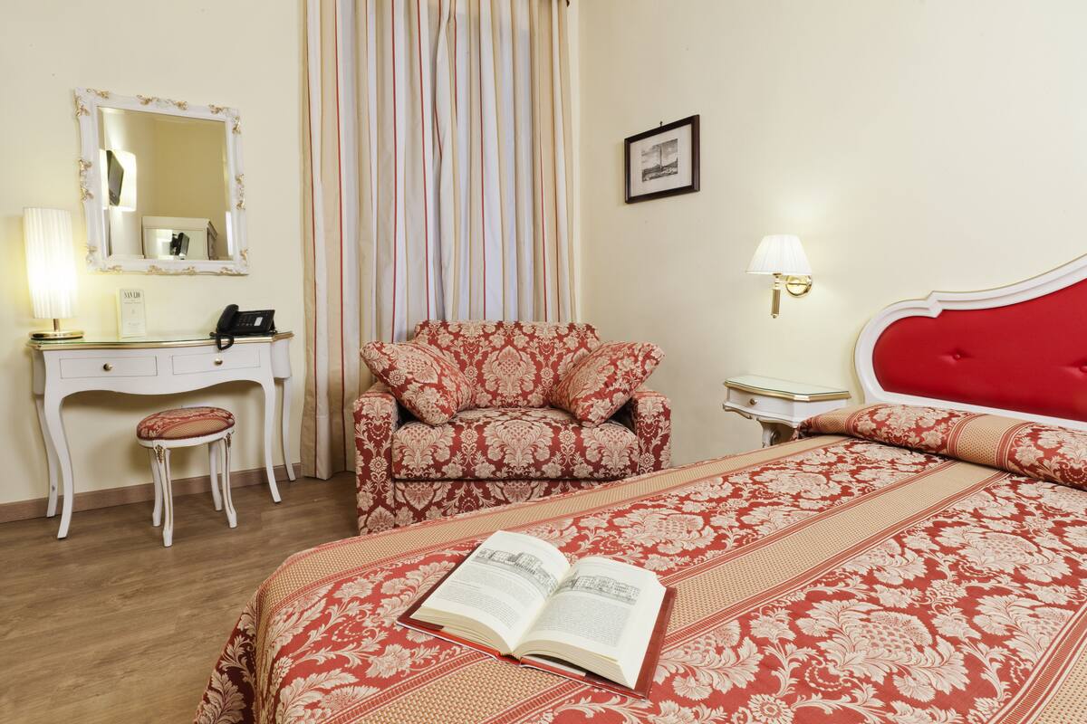 A hotel room with a bed, a desk with a telephone, a chair, a bedside table with a lamp, and a framed picture on the wall.